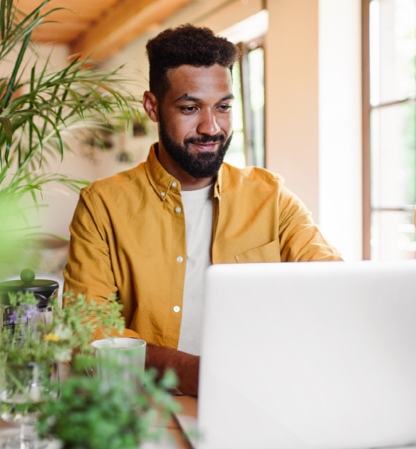 Man sitting at his computer in a yellow shirt surrounded by plants.