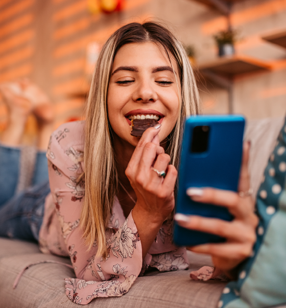 Blonde millennial girl lays on couch eating an Oreo cookie while smiling and looking at a blue mobile phone