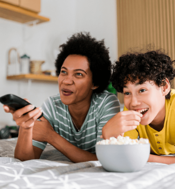 A woman and a young boy sit on a bed, smiling as they watch TV together. The woman holds a remote control, and the boy reaches for popcorn from a bowl in front of them. The setting is cozy and relaxed, with a soft-focus background of a lightly decorated room.