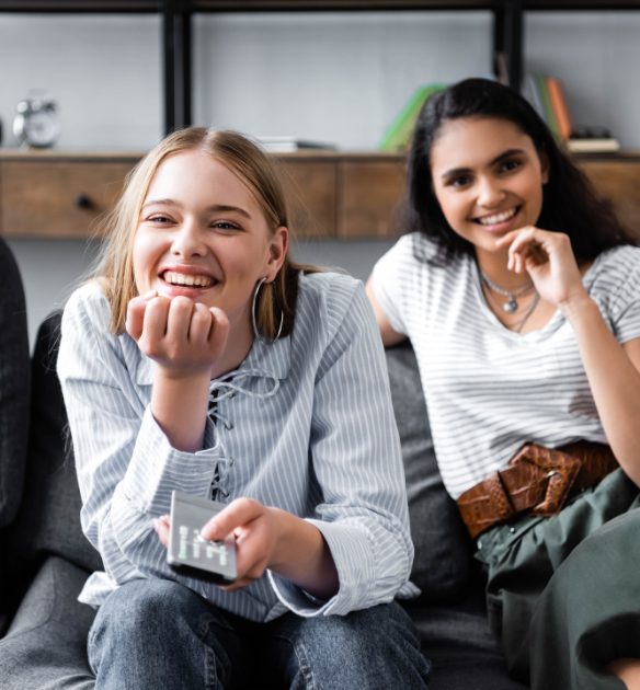 L'image montre deux jeunes femmes souriantes assises sur un canapé dans ce qui semble être un salon. La femme à gauche, vêtue d'une chemise rayée blanche et bleue, tient une télécommande dans sa main droite et s'appuie contre le canapé tout en souriant joyeusement. La femme à droite porte une chemise blanche et un pantalon vert avec une ceinture marron, et est également souriante. En arrière-plan, on peut voir un meuble en bois avec quelques objets de décoration. En haut à gauche de l'image, il y a le logo de « Rakuten Advertising » et le mot « Blog » écrit à côté. L'atmosphère semble conviviale et confortable.