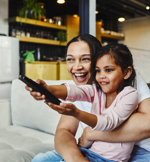 Una mujer y una niña sonriente sentadas en un sofá, sosteniendo un control remoto de televisión y apuntándolo hacia la pantalla en una cálida sala de estar.