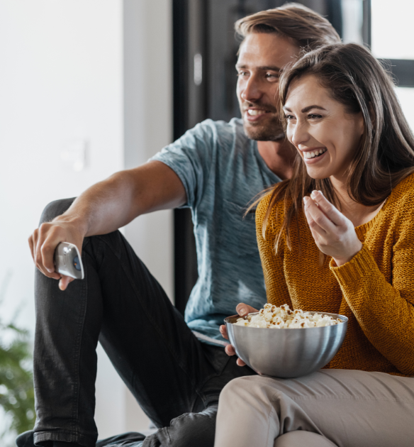 Un homme et une femme sourient tout en regardant la télévision, assis sur un canapé. La femme tient un bol de pop-corn.