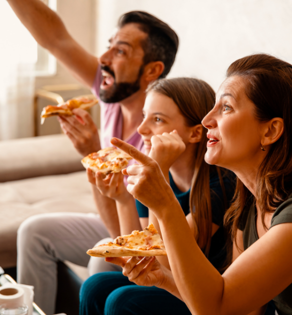 Tre persone sedute su un divano, guardando con entusiasmo qualcosa mentre tengono delle fette di pizza. La persona a sinistra sta esultando, la persona al centro è inclinata in avanti, e la persona a destra sta indicando lo schermo.