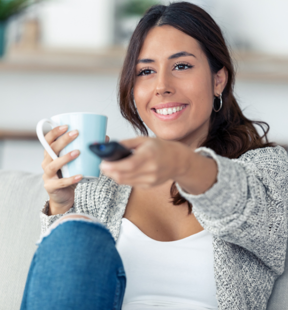 A smiling young woman sits on a couch holding a blue mug and pointing a TV remote control, dressed in a white top, gray knitted cardigan, and blue jeans with a torn knee.