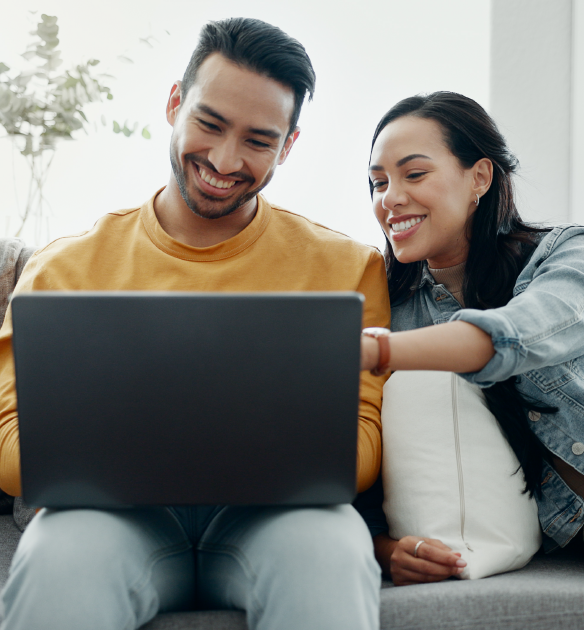 A smiling couple sits close together on a couch, looking at a laptop screen. The man, wearing a mustard-colored sweatshirt, holds the laptop on his lap, while the woman, in a denim jacket, leans in and points at the screen. Both appear to be enjoying themselves as they look at the laptop, possibly engaged in an online activity or watching something entertaining. The background is bright and includes a soft pillow and a partially visible plant.