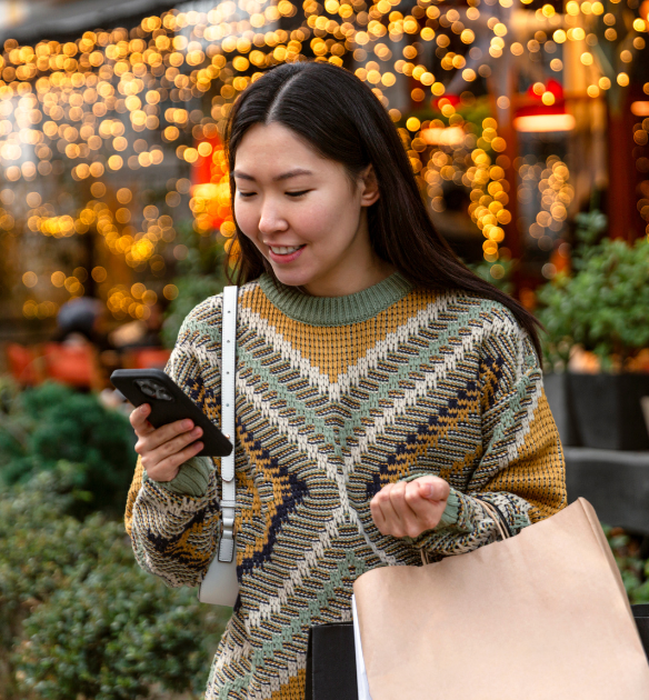 Woman staring down at her phone while smiling in front of christmas lights.